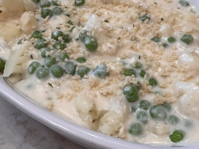 A close-up of a casserole dish filled with cauliflower, green peas, and creamy white sauce, topped with breadcrumbs.