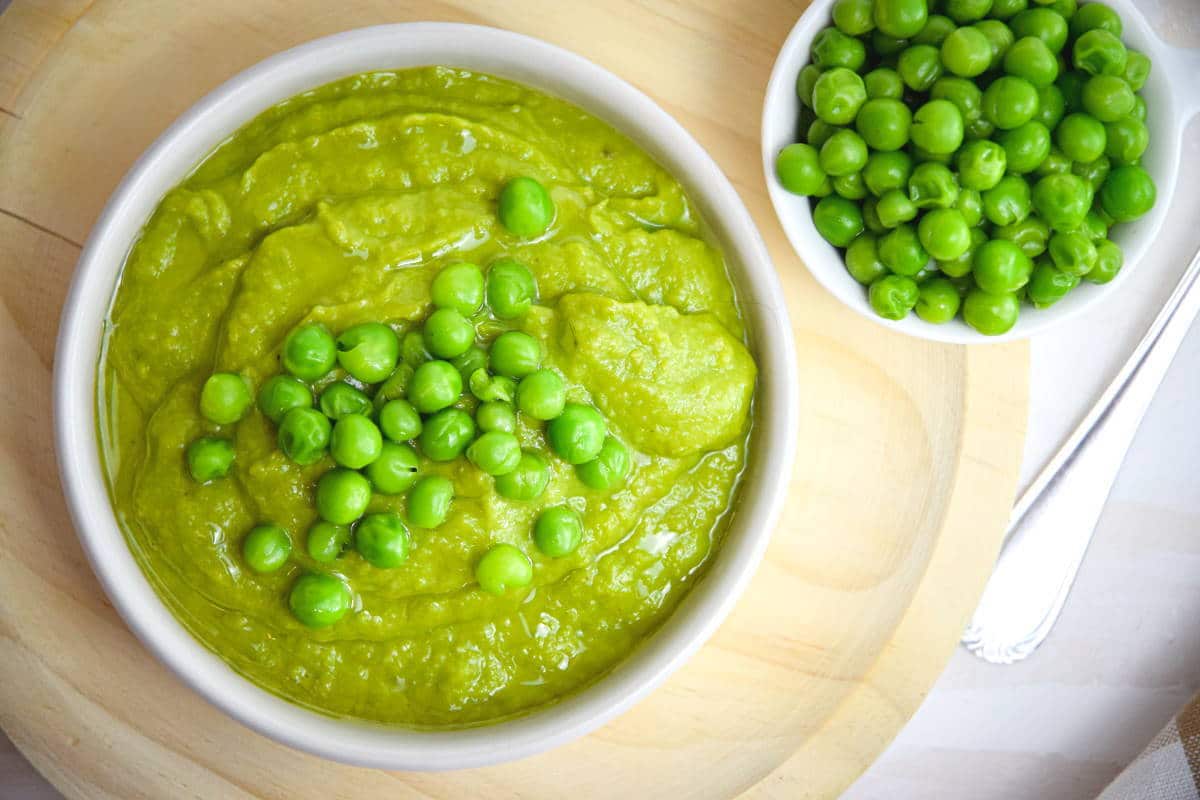 A bowl of mushy peas topped with whole green peas sits next to a small dish of whole green peas on a wooden surface.