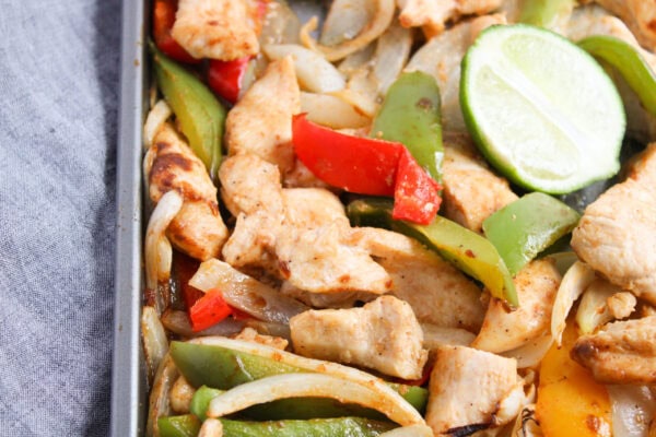 Sheet pan with cooked chicken strips, sliced bell peppers, onions, and two lime slices on a gray surface.