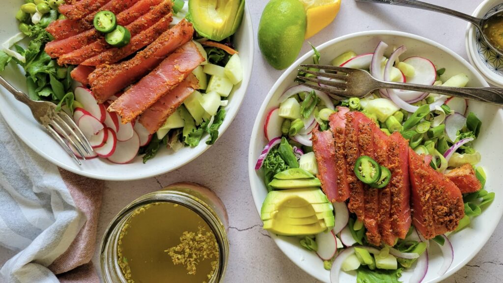 Two bowls of salad with sliced seared tuna, avocado, radish, greens, cucumber, red onion, and a jalapeño slice on top, with a fork and dressing jar nearby.