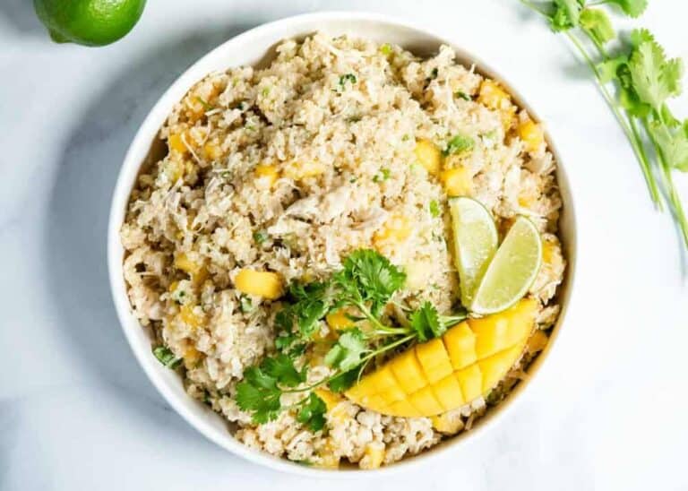 A bowl of quinoa salad with mango chunks, shredded chicken, fresh cilantro, and lime wedges, garnished with a mango slice.