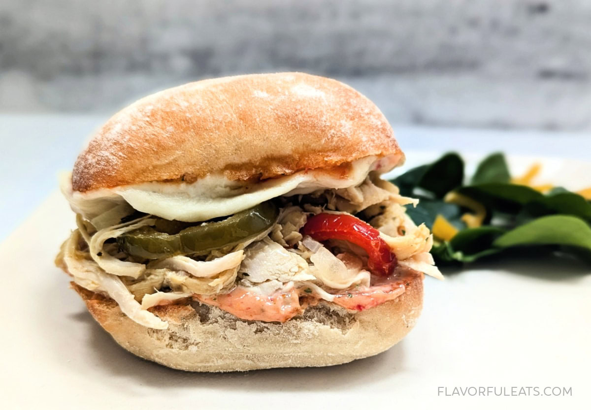 A shredded chicken sandwich with pickles, red peppers, and melted cheese on a rustic bun, served on a white plate with a small side of leafy greens.