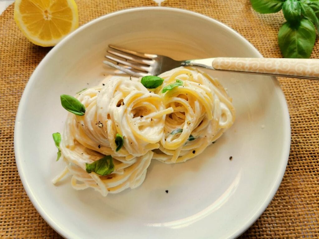 A white plate with two swirls of creamy pasta, garnished with basil leaves and black pepper; a fork, lemon wedge, and basil are also visible.