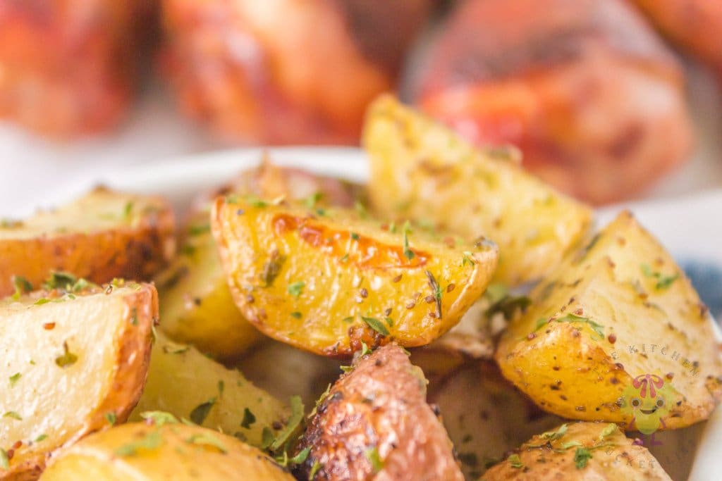 Close-up of roasted potato wedges seasoned with herbs, with blurred baked meat pieces in the background.