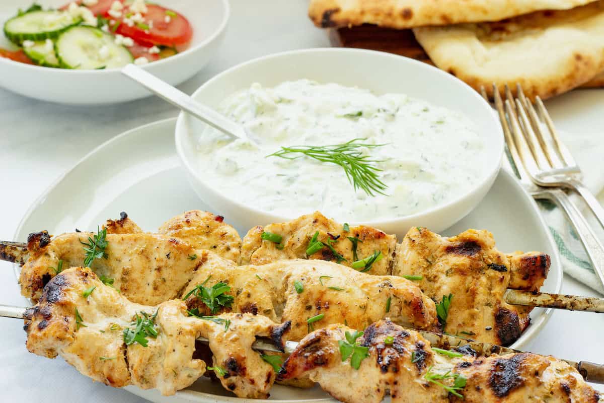 Two grilled chicken skewers garnished with herbs are served on a plate with a bowl of tzatziki sauce, a salad, and pieces of naan bread in the background.
