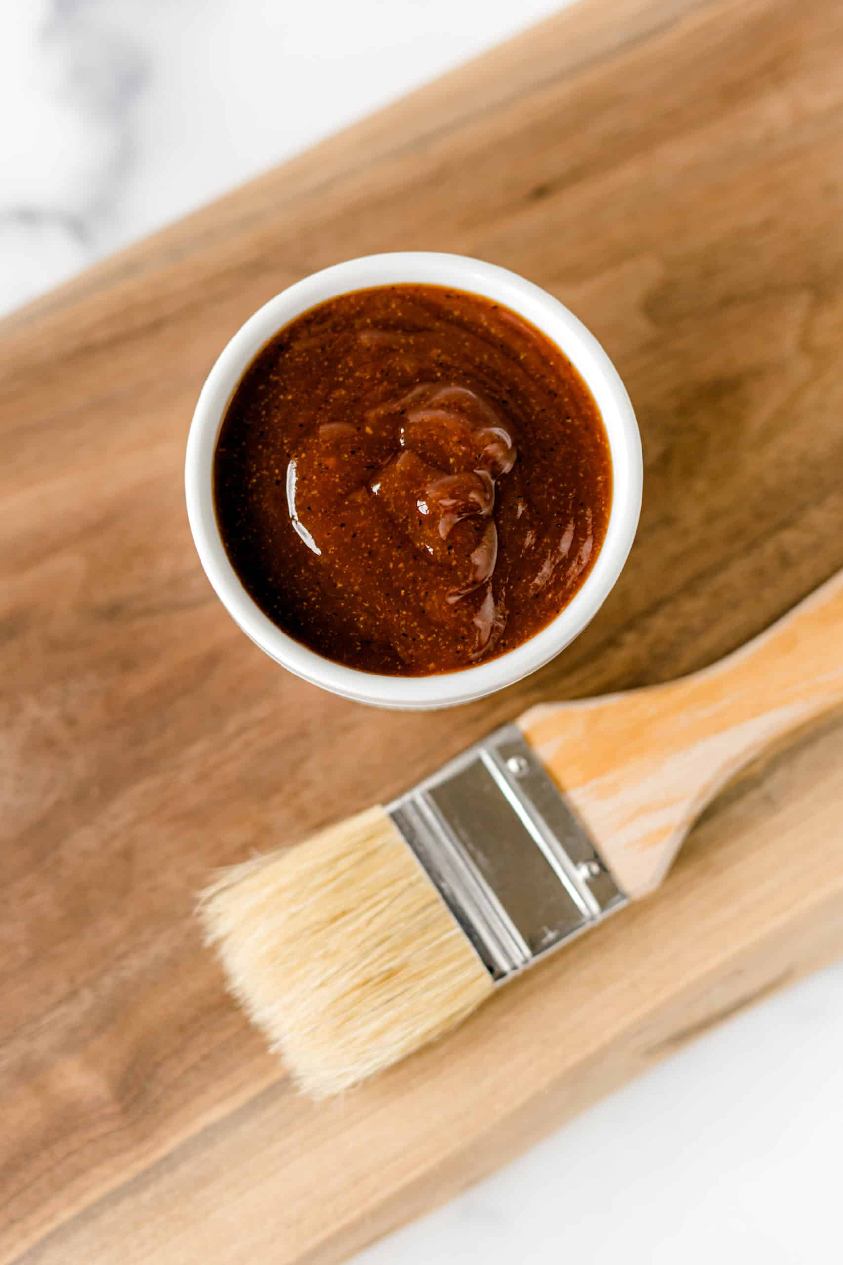 A small white bowl filled with barbecue sauce sits on a wooden board next to a clean basting brush.