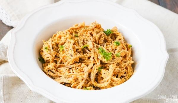 A white bowl filled with shredded chicken mixed with spices and garnished with fresh herbs, placed on a light-colored cloth.