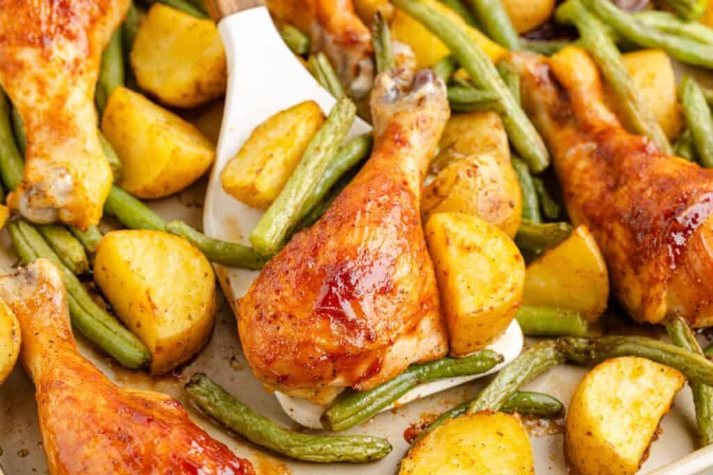 Baked chicken drumsticks, green beans, and roasted potato pieces on a baking sheet, with a spatula lifting one drumstick and some vegetables.