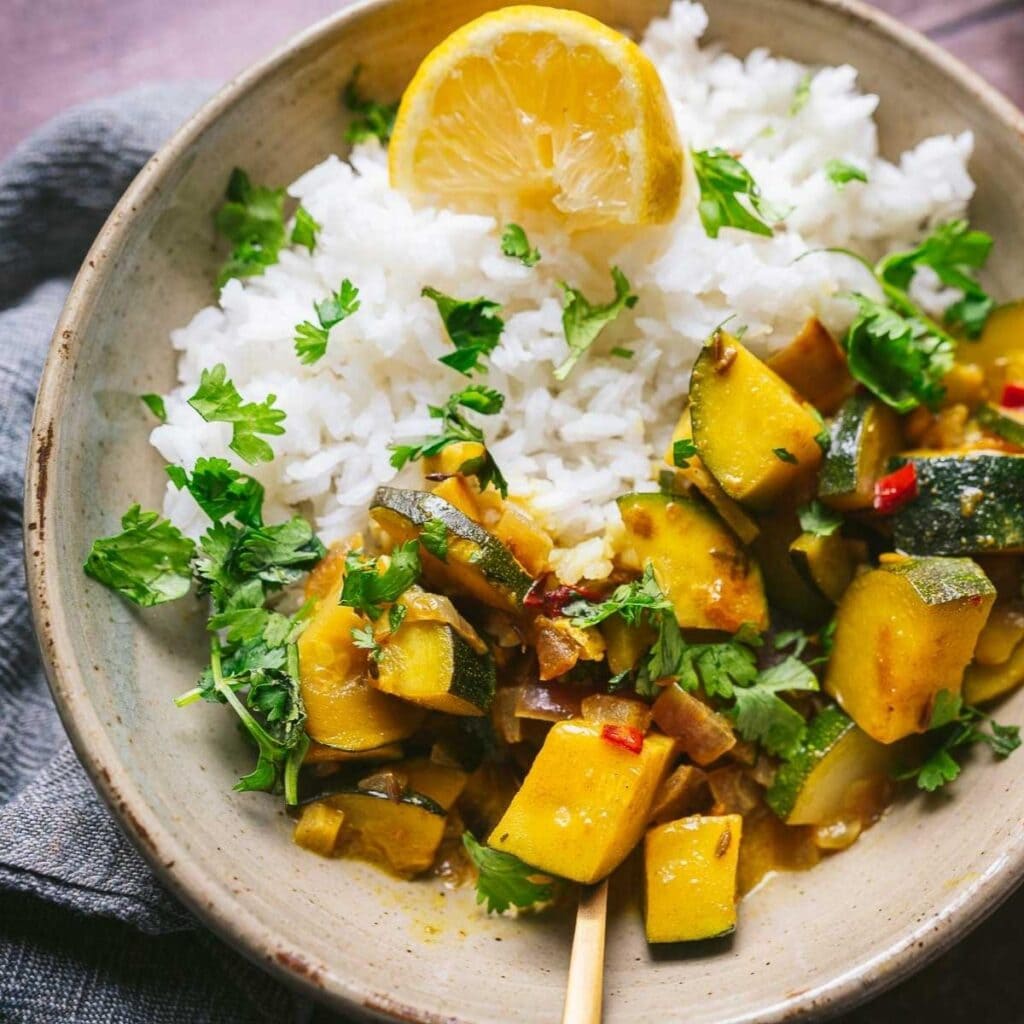 A bowl of white rice with curried zucchini, garnished with chopped cilantro and a lemon wedge.