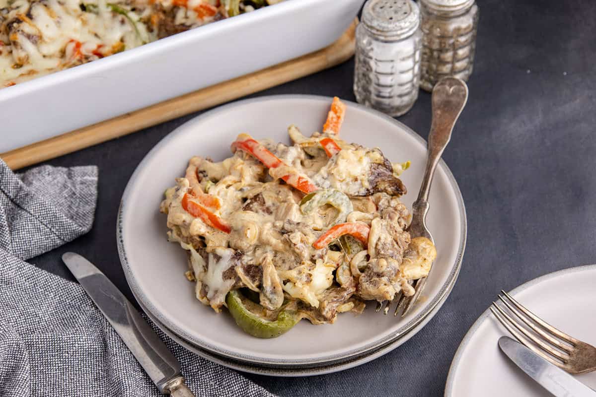A serving of cheesy casserole with sliced beef, red and green bell peppers on a plate, with a fork beside salt and pepper shakers.