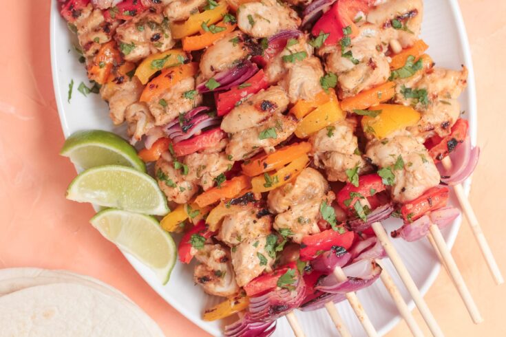 Grilled chicken and vegetable skewers garnished with chopped herbs, served on a white plate with lime wedges on the side.
