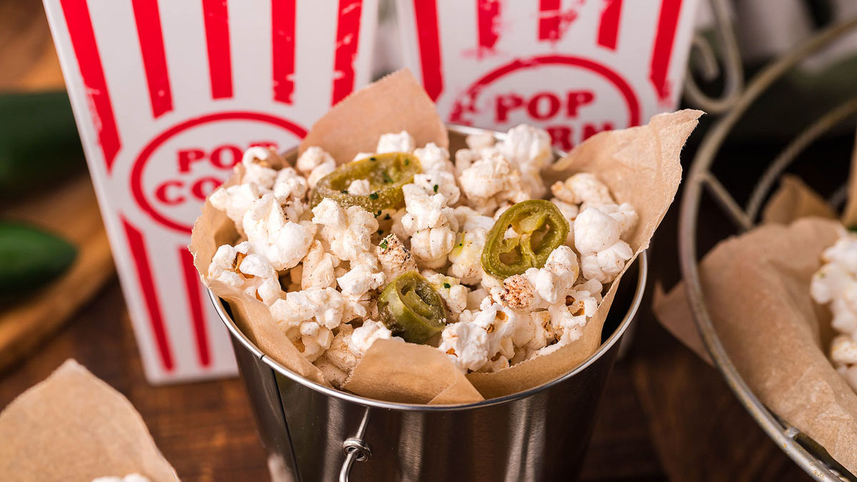 A metal bucket lined with brown paper is filled with popcorn topped with sliced jalapeรฑos, with striped popcorn containers in the background.
