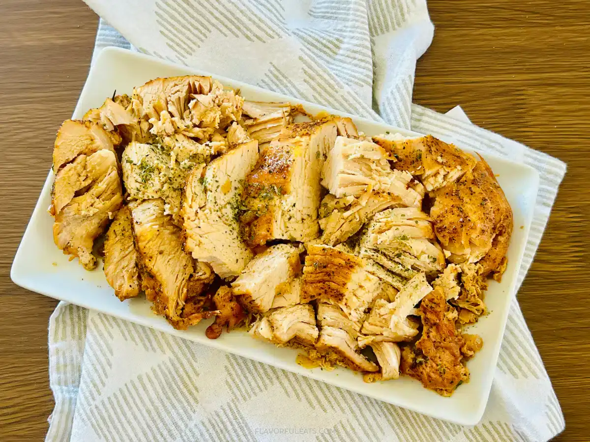 Sliced cooked turkey breast seasoned with herbs, arranged on a rectangular white plate, placed on a patterned cloth atop a wooden surface.