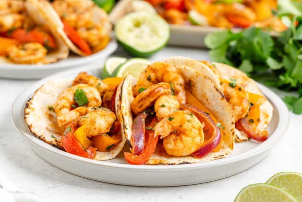 Three shrimp fajita tacos with sautéed shrimp, red and yellow bell peppers, and onions, served on a white plate with lime wedges and fresh cilantro in the background.