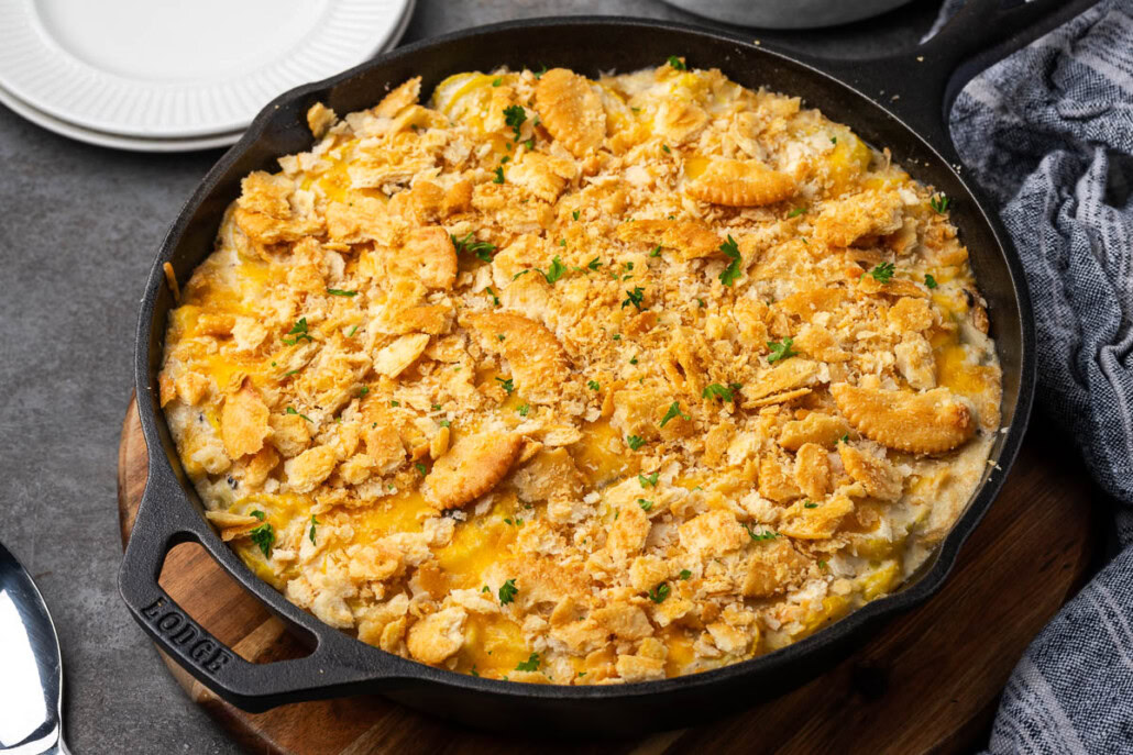 A cast iron skillet filled with a baked casserole topped with crushed crackers and garnished with chopped herbs, placed on a wooden board.