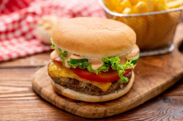 A cheeseburger with lettuce, tomato, sauce, and a beef patty on a wooden board, with a basket of French fries in the background.