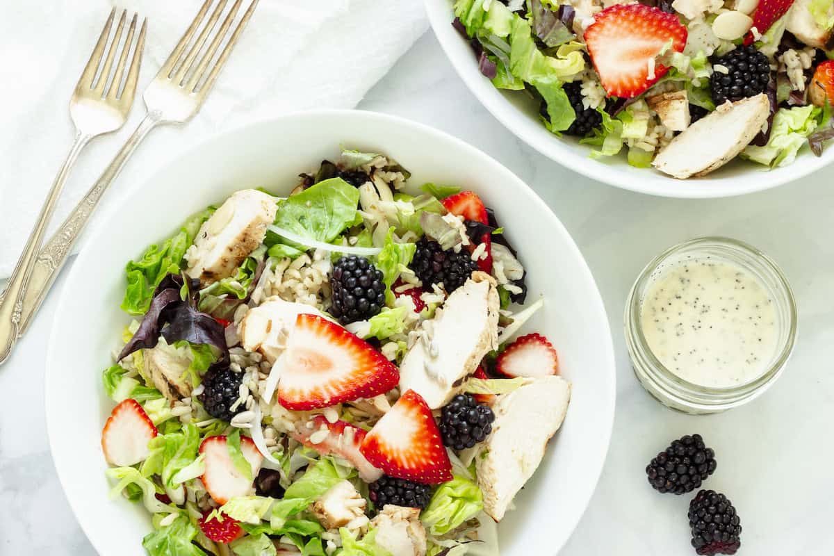 A white bowl filled with salad, grilled chicken slices, strawberries, blackberries, and greens, with a jar of dressing and two forks nearby.