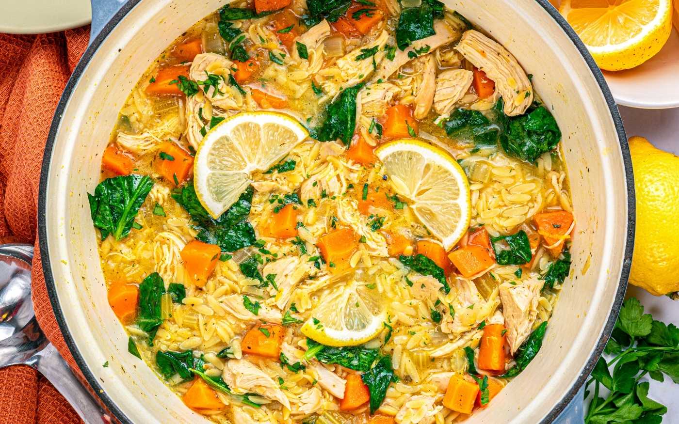 A pot of chicken soup with shredded chicken, orzo, spinach, carrots, herbs, and lemon slices, viewed from above.