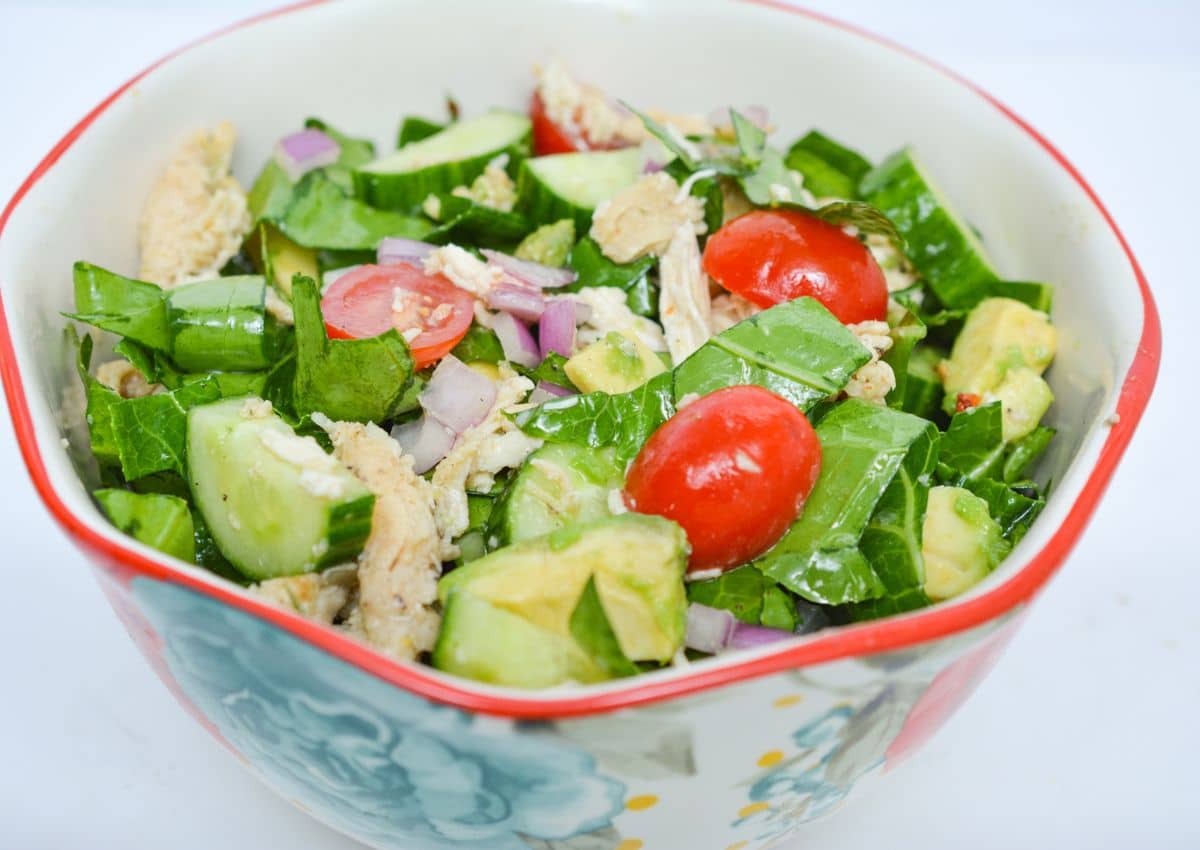 A bowl of salad containing chopped spinach, avocado, cucumber, grape tomatoes, red onion, and pieces of cooked chicken.