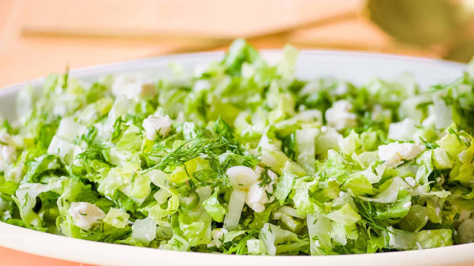 A close-up of a fresh green salad with chopped lettuce, herbs, and small pieces of white cheese on a white plate.