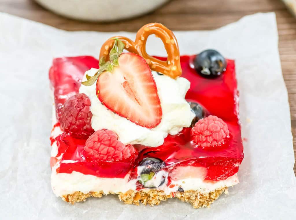 A dessert bar with a pretzel crust, creamy layer, red gelatin, berries, whipped cream, and garnished with a strawberry slice, raspberries, blueberries, and a pretzel twist.