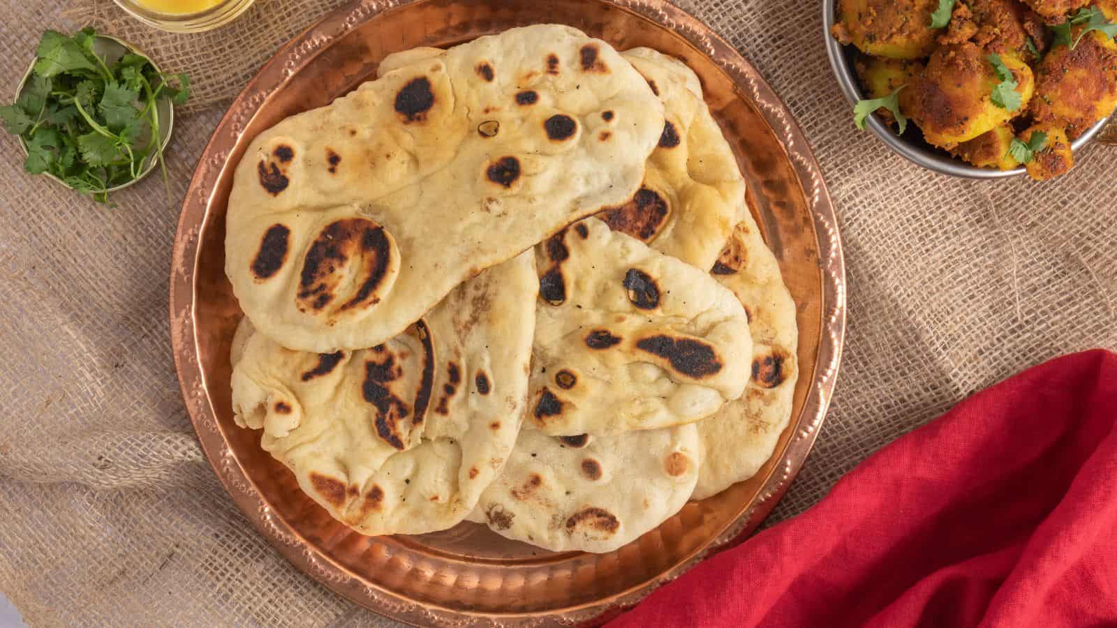 A copper plate with several pieces of naan bread, a bowl of cooked vegetables, a sprig of cilantro, and a red cloth on a burlap surface.