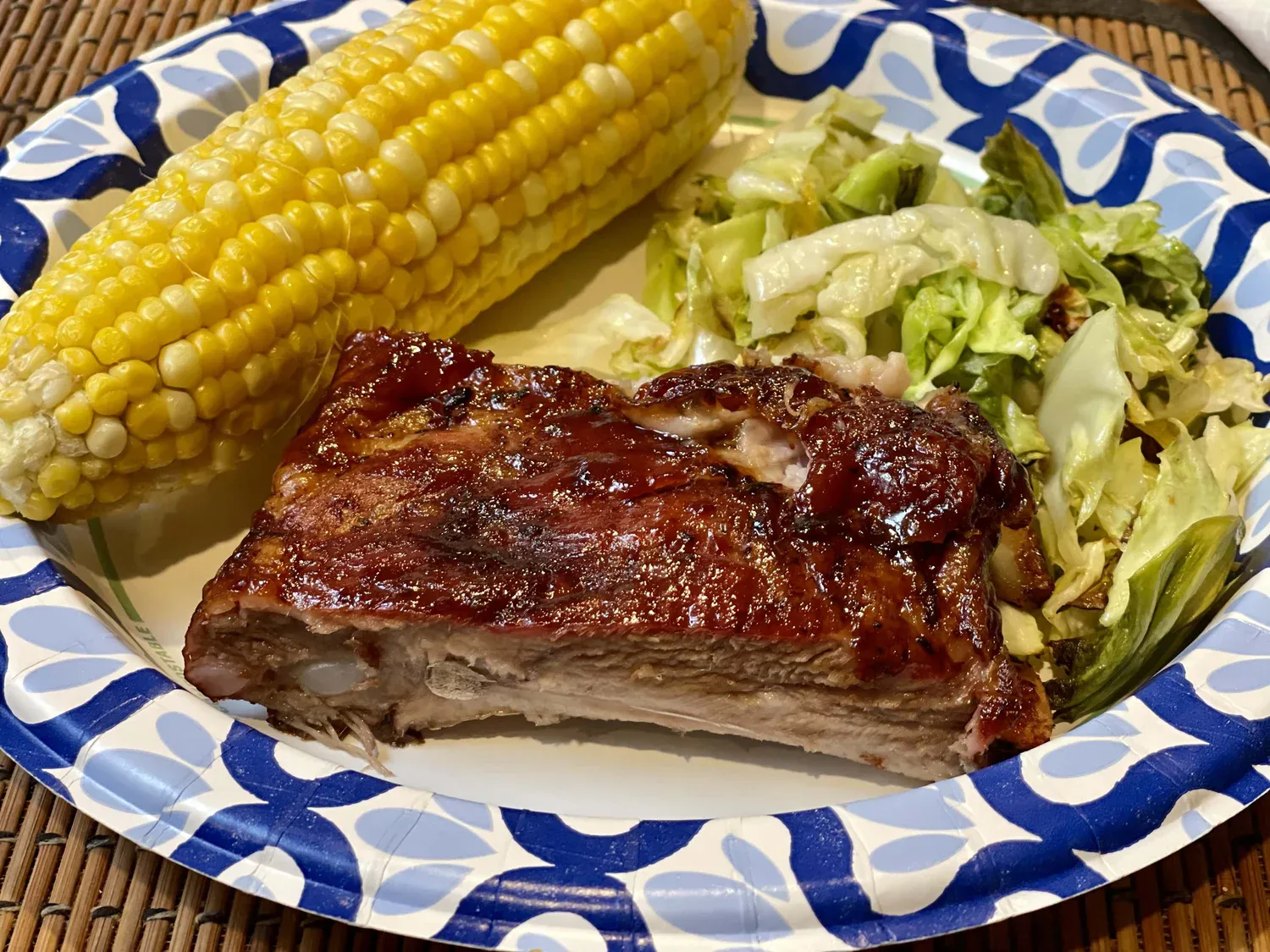 A paper plate with a serving of barbecued ribs, corn on the cob, and cooked cabbage.