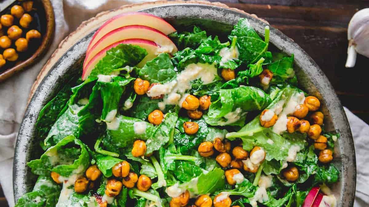 A bowl of green leafy salad topped with roasted chickpeas, apple slices, and creamy dressing.