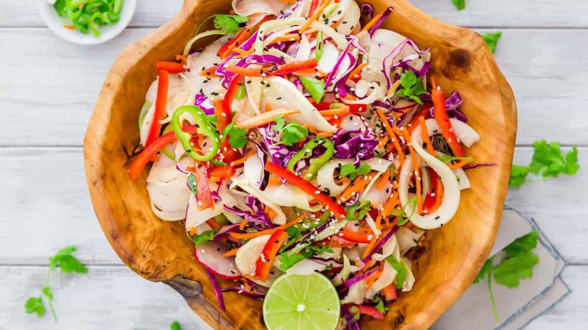 A wooden bowl filled with a colorful salad of shredded cabbage, carrots, red bell peppers, onions, cilantro, and black sesame seeds, garnished with a lime slice.