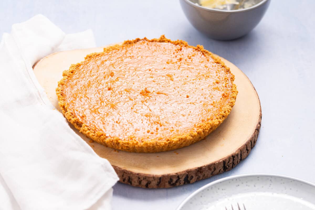 A baked custard pie sits on a round wooden board next to a white cloth, with a bowl and an empty plate nearby.