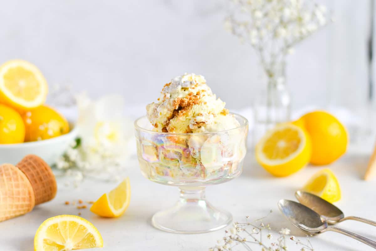 A glass dish of lemon ice cream with a crumb topping sits on a white surface, surrounded by lemon halves, ice cream cones, and metal spoons.