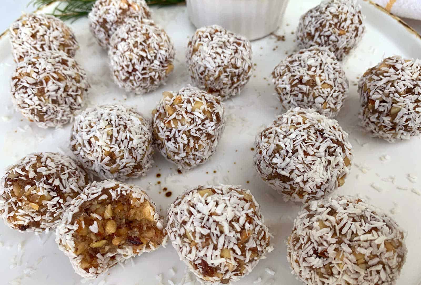 A plate of round energy bites coated in shredded coconut, with one showing the nut and fruit mixture inside.