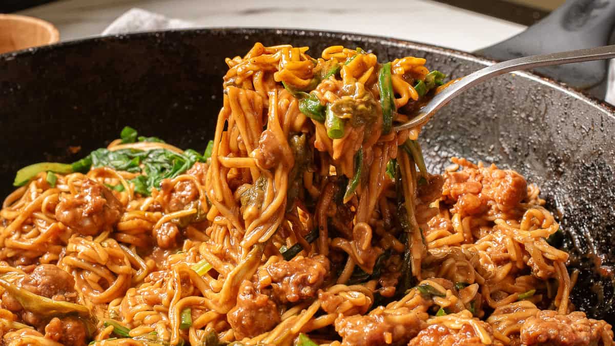 A close-up of cooked noodles with ground meat and spinach being lifted from a pan with a spoon.