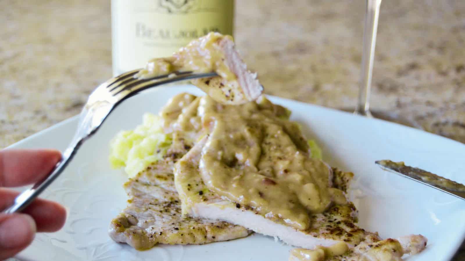 A close-up of a fork holding a slice of pork chop topped with creamy mushroom sauce, with more pork and sauce on a white plate. A wine bottle is blurred in the background.