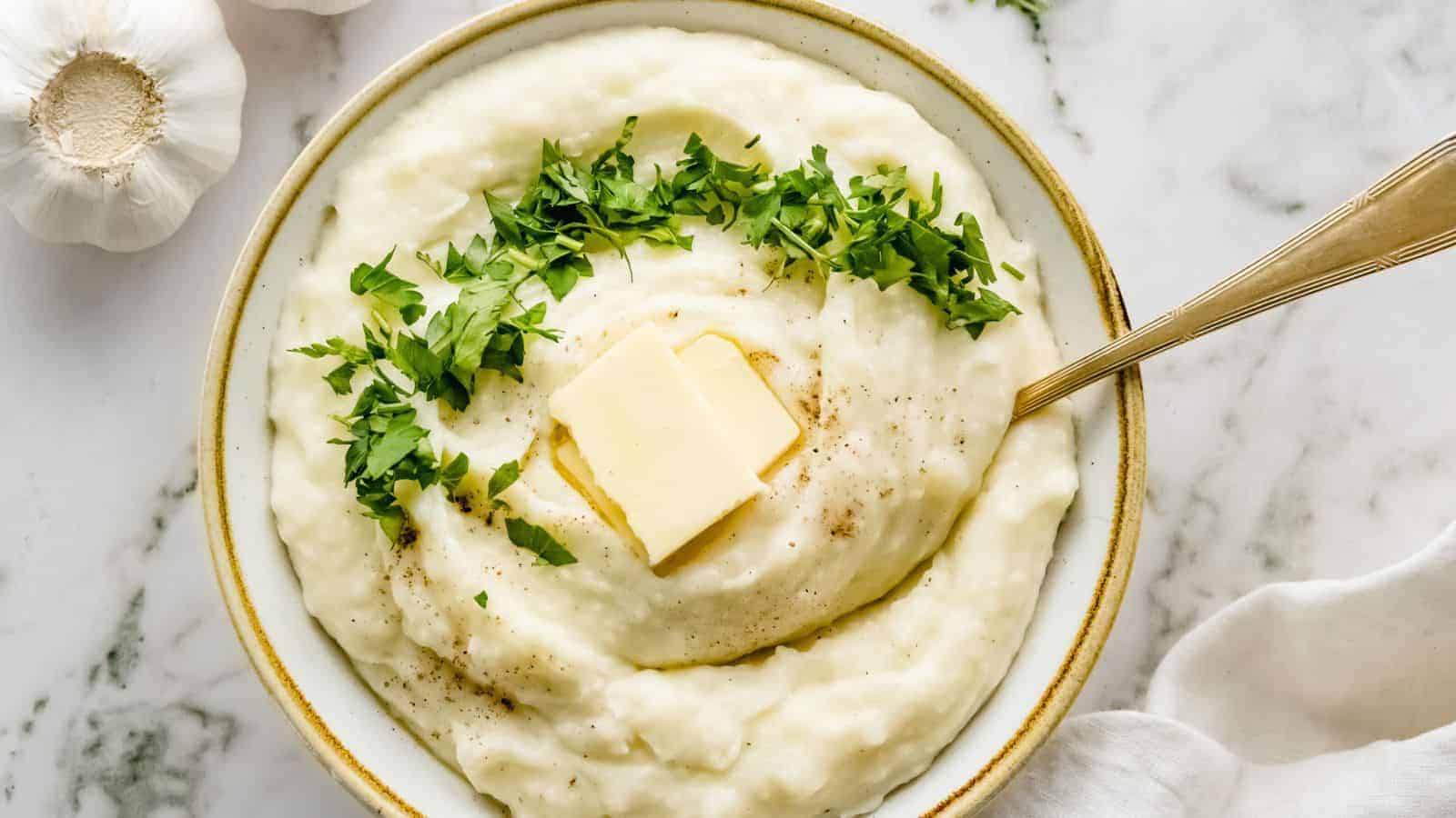 Bowl of mashed potatoes topped with chopped parsley and two pats of butter, with a spoon resting inside, on a marble surface.