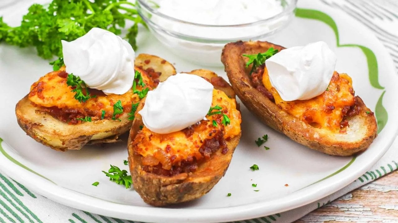 Three loaded potato skins topped with melted cheese, bacon bits, chopped parsley, and dollops of sour cream on a white plate. A bowl of sour cream and parsley garnish are in the background.