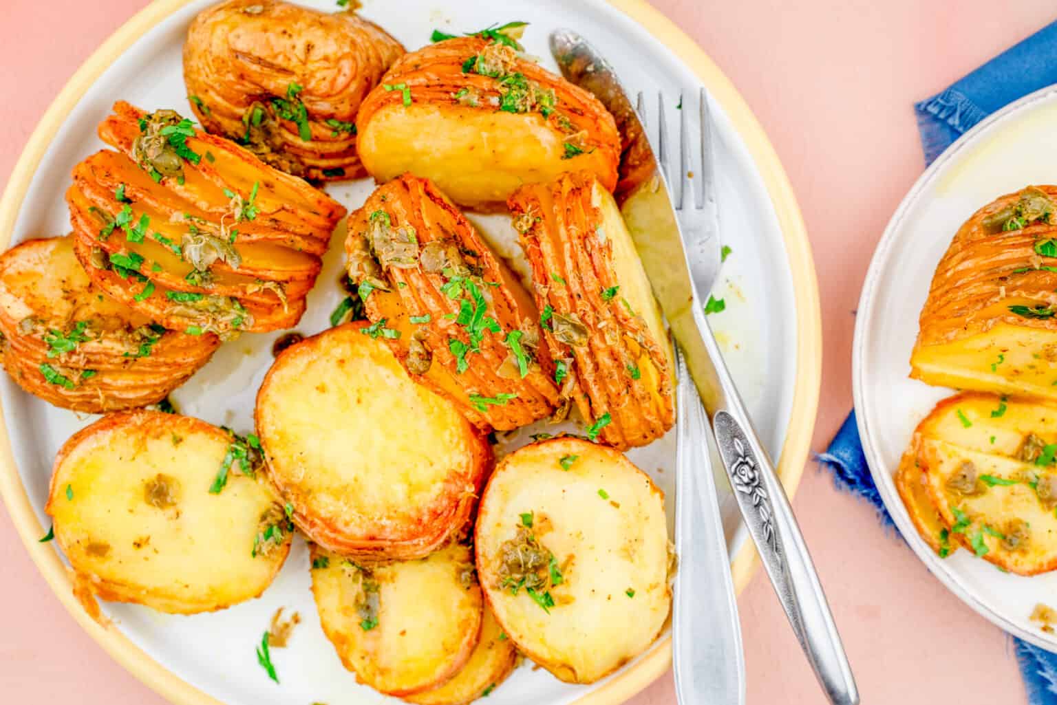 A plate of sliced, roasted hasselback potatoes garnished with herbs, with a fork and knife resting on the plate.