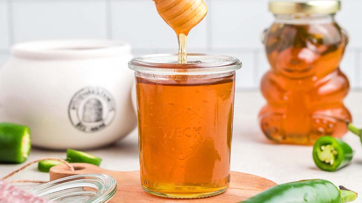 A jar of honey with honey being drizzled in, surrounded by a honey bear jar, a ceramic container, and sliced green jalapeรฑos on a wooden board.