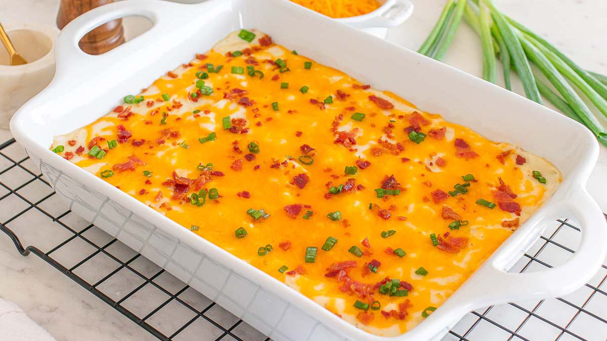 A baked casserole topped with melted cheddar cheese, chopped bacon, and sliced green onions in a white dish on a cooling rack.