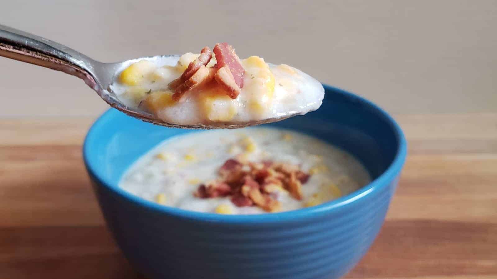 A spoonful of creamy soup with corn and bacon held above a blue bowl filled with the same soup, resting on a wooden surface.