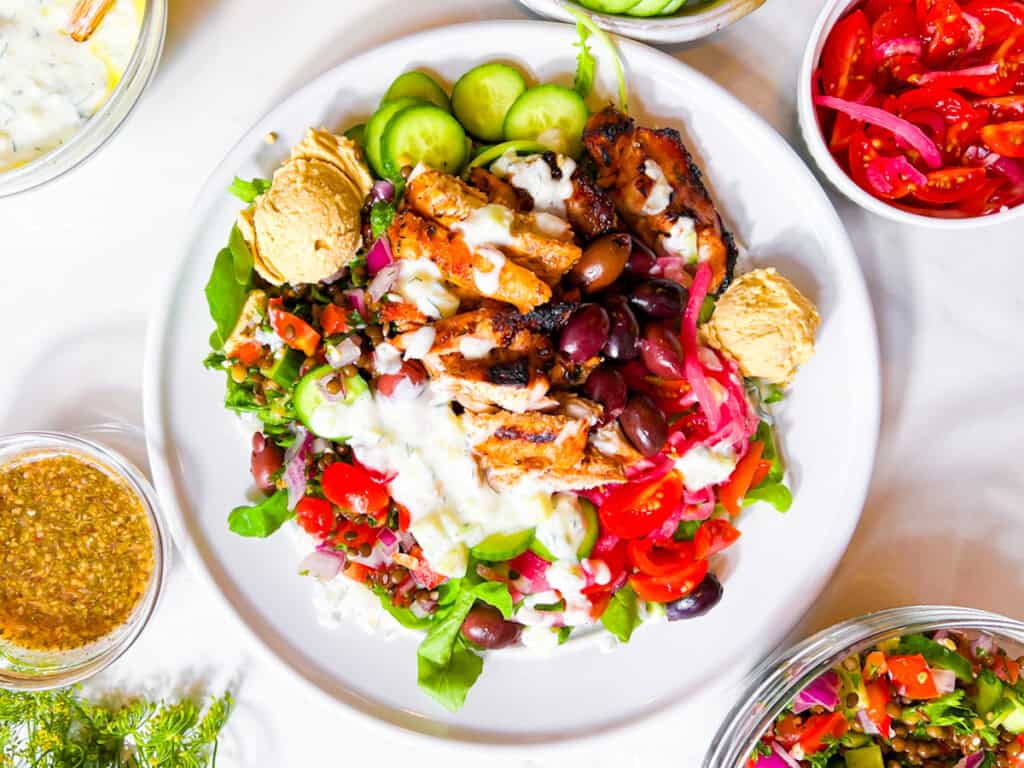 A plate of Mediterranean salad with grilled chicken, hummus, cucumbers, olives, greens, tomatoes, and creamy dressing, surrounded by small bowls of vegetables and sauce.