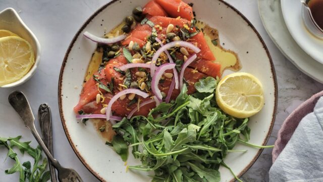 A plate with smoked salmon topped with red onion, capers, and herbs, served with a side of arugula and a halved lemon.