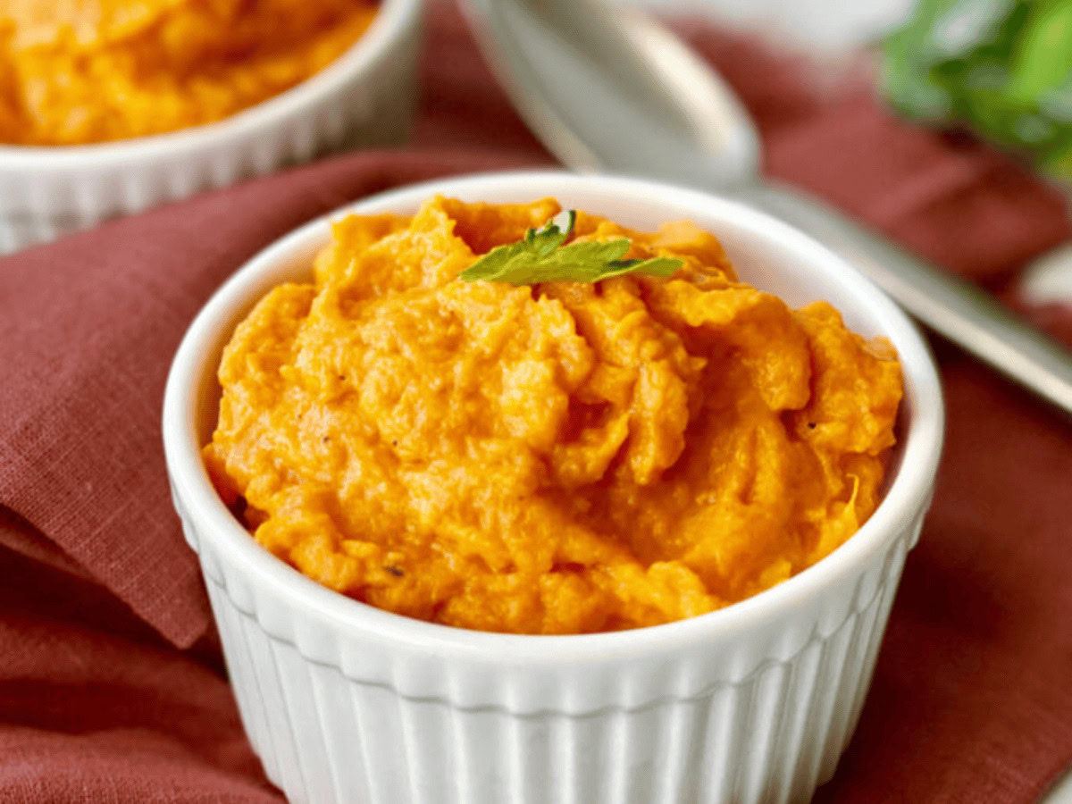 A white ramekin filled with orange sweet potato mash, garnished with a small green herb, placed on a red cloth with a spoon in the background.
