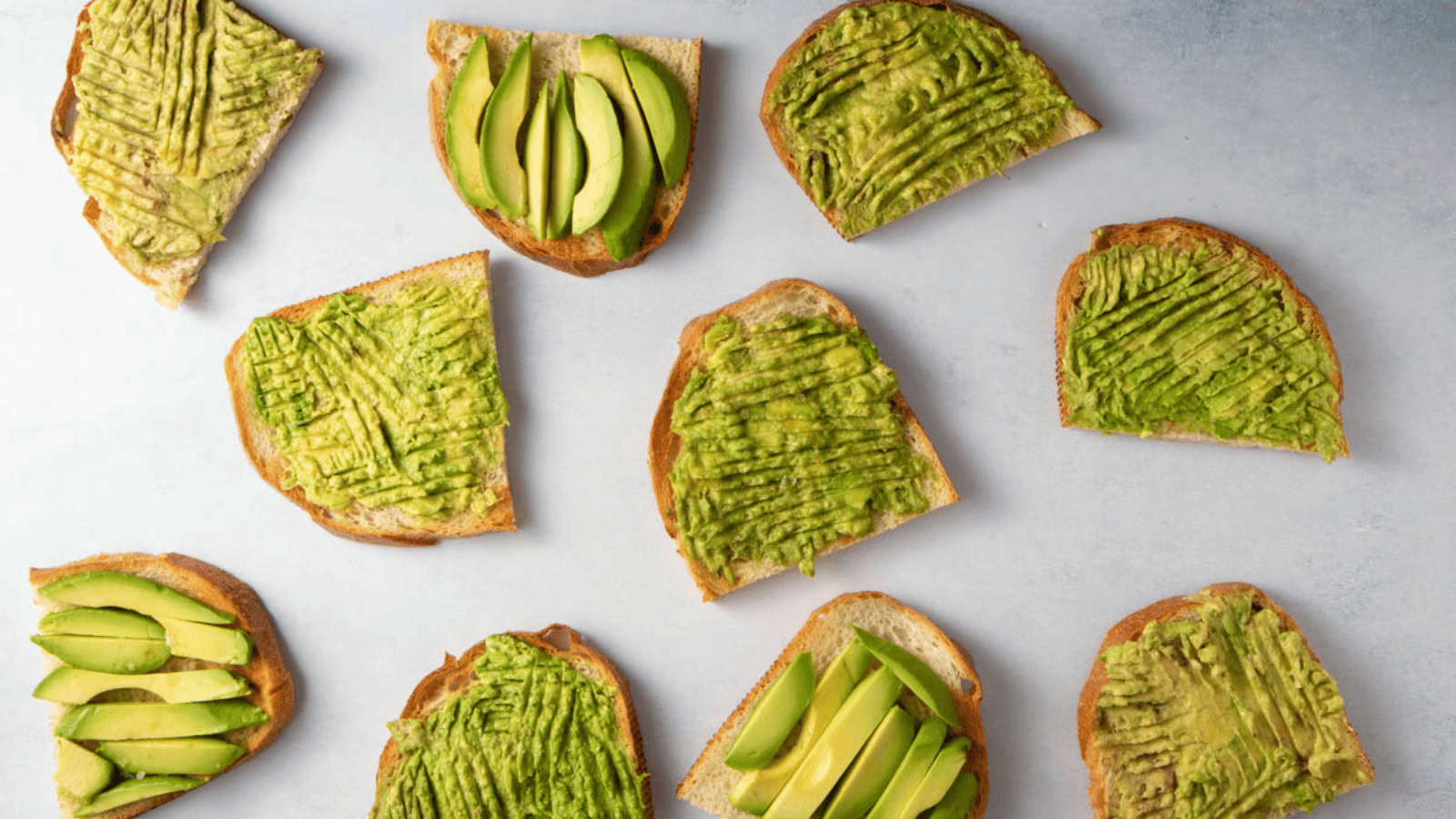 Eight slices of toast topped with mashed or sliced avocado are arranged on a light-colored surface.
