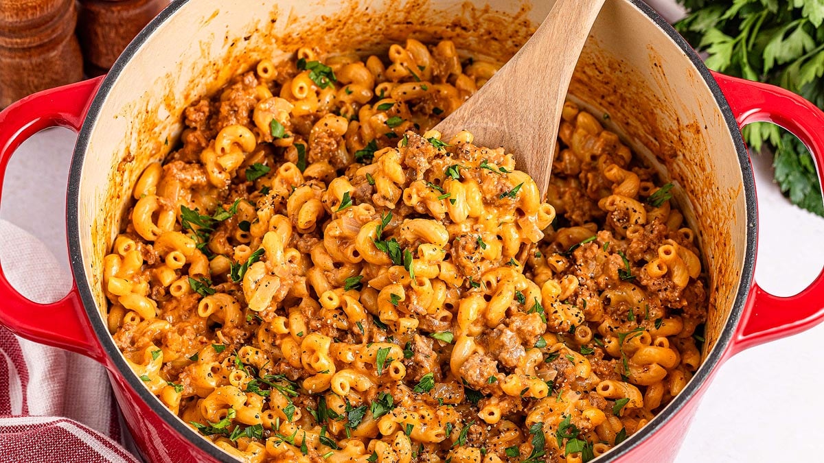 A red pot filled with cooked macaroni mixed with ground meat and sauce, garnished with chopped parsley, with a wooden spoon resting inside.