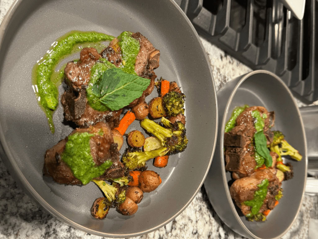 Two gray bowls containing grilled lamb chops topped with green sauce and mint, served with roasted carrots, broccoli, and baby potatoes on a granite countertop.