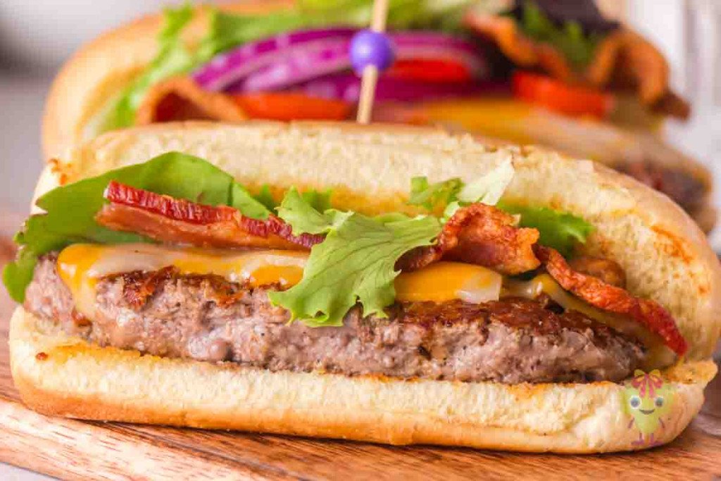 A close-up of a sandwich with a grilled beef patty, cheddar cheese, crispy bacon, lettuce, and a hoagie bun, with another similar sandwich in the background.