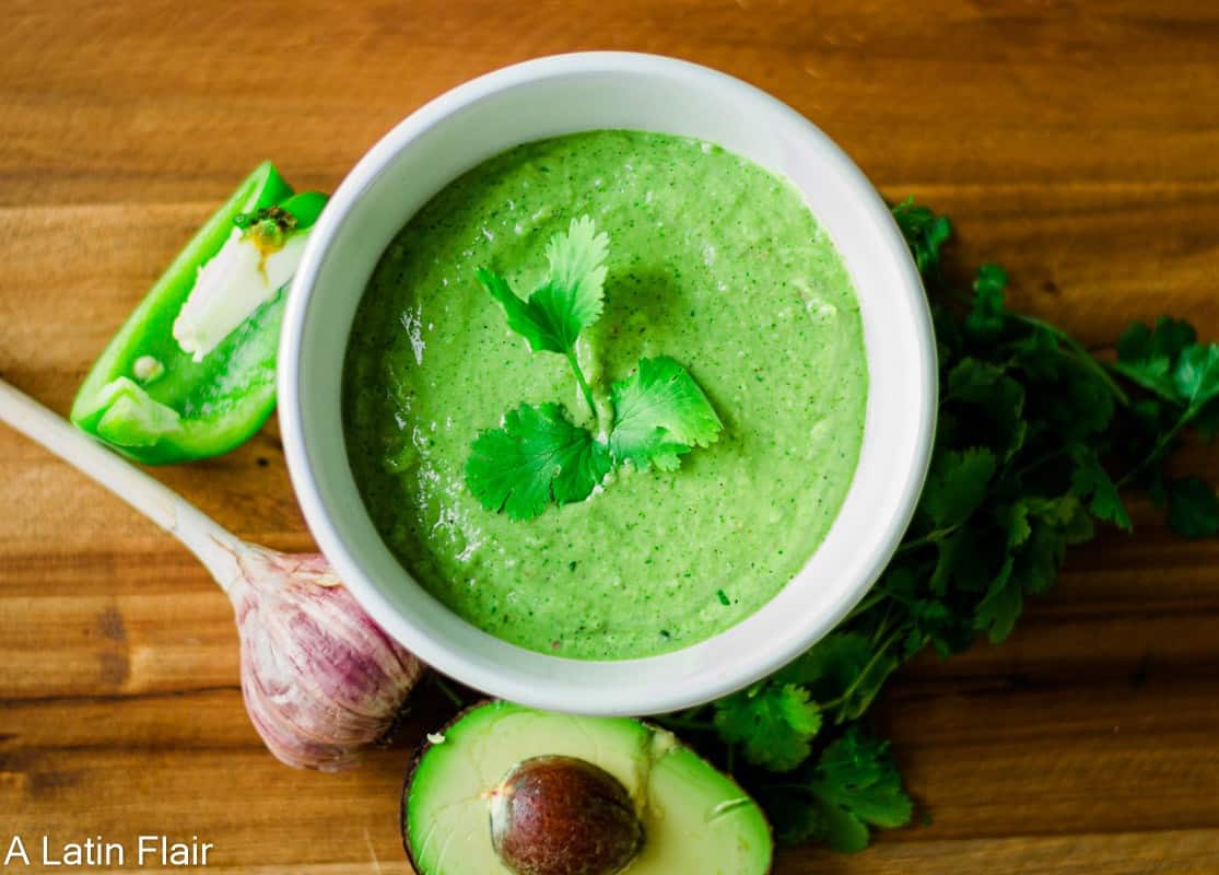 A bowl of green sauce garnished with cilantro, surrounded by fresh cilantro, garlic, jalapeño, and half an avocado on a wooden surface.