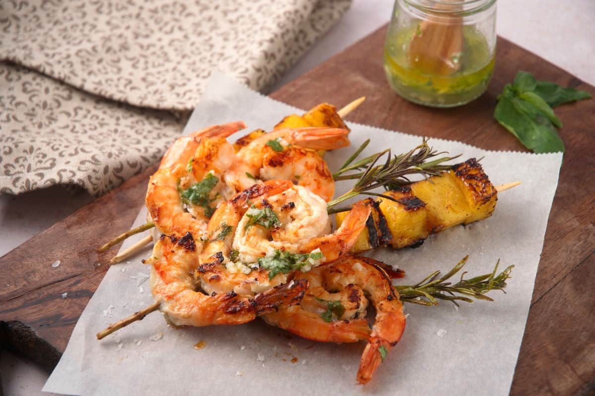 Grilled shrimp skewers with herbs are served on parchment paper beside grilled pineapple and a small jar of green sauce on a wooden board.