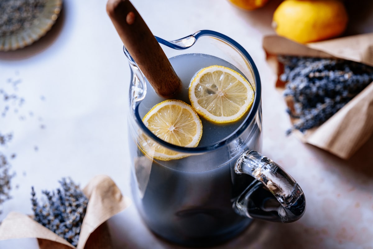 A glass pitcher filled with a dark purple drink, garnished with two lemon slices and a wooden stirrer, surrounded by dried lavender and lemons.